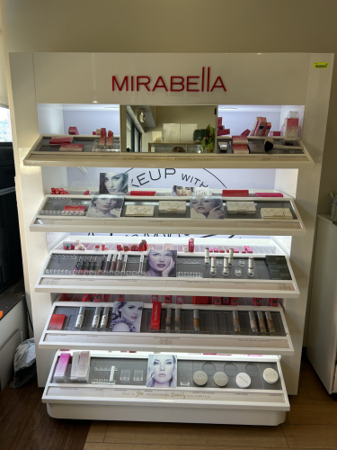 MIRABELLA LIGHTED RETAIL DISPLAY SHELF WITH MAKEUP CONTENTS. INCLUDES MULTIPLE TIERS WITH ASSORTED COSMETICS SUCH AS LIPSTICKS, GLOSSES, FOUNDATIONS, POWDERS, BLUSHES, AND TESTER UNITS. BRANDED SIGNAGE AND BUILT-IN LIGHTING. CONTENTS INCLUDED AS SHOWN.