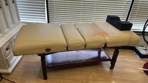 ADJUSTABLE MASSAGE TABLE WITH TAN VINYL UPHOLSTERY AND WOOD FRAME. FEATURES MULTI-SECTION PAD, FACE CRADLE, SIDE ARM SUPPORTS, AND LOWER STORAGE SHELF