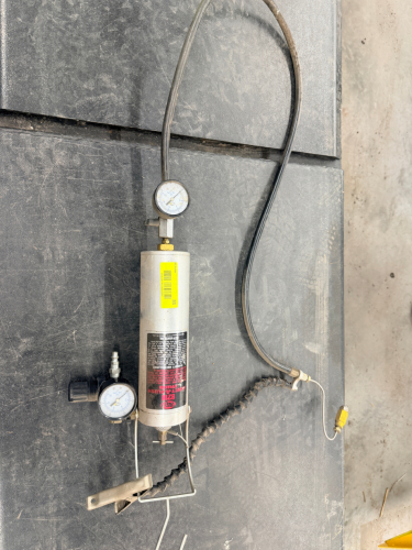 PNEUMATIC BRAKE BLEEDER UNIT WITH PRESSURE GAUGE, HOSE, AND METAL RESERVOIR CANISTER. INCLUDES HAND TRIGGER VALVE AND HANGING HOOK BRACKET.