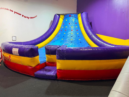 COMMERCIAL INFLATABLE SLIPPERY SLOPE SLIDE WITH PADDED ENTRY, HIGH SIDEWALLS, AND ENCLOSED LANDING AREA. UNIT FEATURES MULTI-COLOR VINYL CONSTRUCTION WITH TOP SAFETY NETTING.