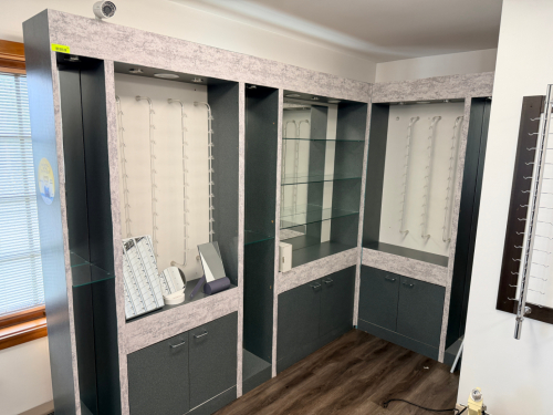 MODULAR EYEGLASS DISPLAY WALL SYSTEM WITH MULTIPLE DISPLAY BAYS, GLASS SHELVING, LOWER STORAGE CABINETS, AND INTEGRATED LIGHTING. FEATURES LAMINATE WOOD CONSTRUCTION WITH BUILT-IN MIRRORS, METAL EYEGLASS DISPLAY RODS, AND CORNER CONFIGURATION