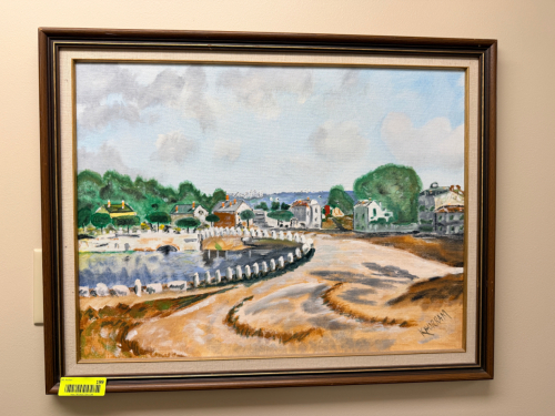 FRAMED IMPRESSIONIST-STYLE LANDSCAPE OIL PAINTING ON CANVAS DEPICTING A RIVERSIDE OR COASTAL VILLAGE SCENE WITH ROADWAY, WATER, AND HOUSES.
