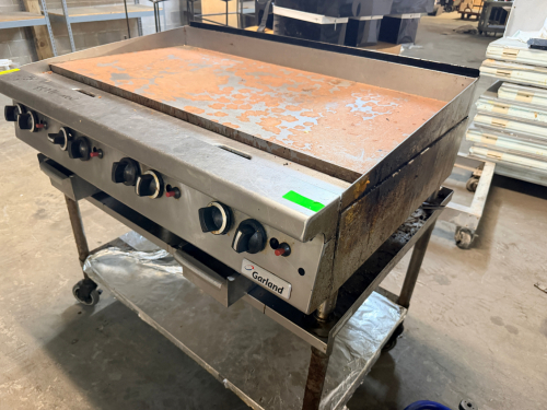 GARLAND COMMERCIAL GAS FLAT-TOP GRIDDLE WITH LARGE STEEL COOKING SURFACE, SIX FRONT-MOUNTED CONTROL KNOBS, MANUAL IGNITION CONTROLS, STAINLESS STEEL FRONT AND SIDES, SPLASH GUARDS, AND FRONT GREASE DRAWERS.
