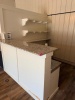 CUSTOM BUILT L SHAPED SERVICE COUNTER WITH POLISHED GRANITE COUNTERTOP, RAISED FRONT AND SIDE PANELS, LOWER GRANITE SERVICE LEDGE, AND THREE WALL-MOUNTED DISPLAY SHELVES WITH DECORATIVE BRACKETS. WHITE PAINTED WOOD CONSTRUCTION WITH LIGHT-COLORED SPECKLED - 2