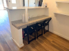 CUSTOM BUILT L SHAPED SERVICE COUNTER WITH POLISHED GRANITE COUNTERTOP, RAISED FRONT AND SIDE PANELS, LOWER GRANITE SERVICE LEDGE, AND THREE WALL-MOUNTED DISPLAY SHELVES WITH DECORATIVE BRACKETS. WHITE PAINTED WOOD CONSTRUCTION WITH LIGHT-COLORED SPECKLED - 3