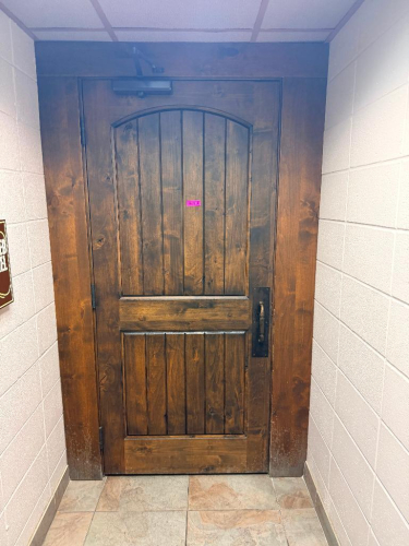 SOLID CORE WOOD DOOR WITH STAINED FINISH, ARCHED RAISED PANEL DESIGN, HEAVY-DUTY HINGES, METAL PULL HANDLE, AND OVERHEAD HYDRAULIC DOOR CLOSER.