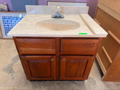 WOOD BATHROOM VANITY WITH MEDIUM BROWN FINISH, RAISED-PANEL CABINET DOORS, TWO FALSE DRAWER FRONTS, CULTURED MARBLE-LOOK VANITY TOP WITH INTEGRATED OVAL SINK, BACKSPLASH, AND SINGLE-HANDLE FAUCET.