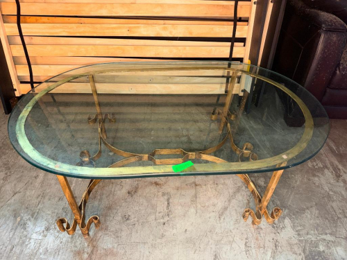 OVAL GLASS-TOP COFFEE TABLE WITH DECORATIVE SCROLLED METAL BASE, GOLD-TONE FINISH, CURVED LEGS, AND BEVELED-EDGE GLASS TOP.