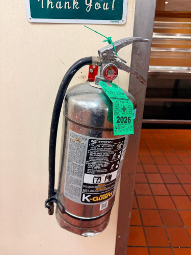 K-GUARD WET CHEMICAL CLASS K FIRE EXTINGUISHER WITH STAINLESS STEEL CYLINDER, DISCHARGE HOSE, PRESSURE GAUGE, SAFETY PIN AND WALL-MOUNT BRACKET. DESIGNED FOR CLASS K COOKING OIL AND GREASE FIRES. INSPECTION TAG VISIBLE AND MARKED 2026.