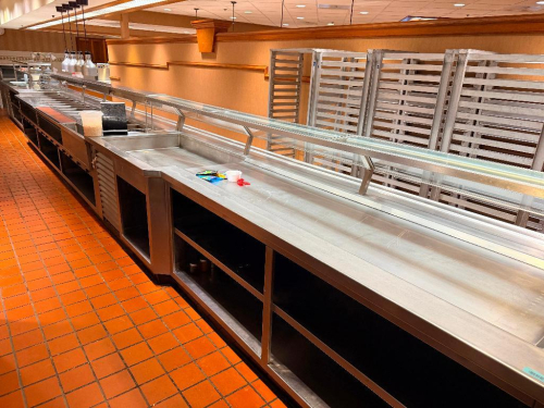 APPROX 50' X 25' &ldquo;L&rdquo; SHAPED COMMERCIAL STAINLESS STEEL BUFFET / SERVING LINE WITH CHECKOUT COUNTER, MULTIPLE INTEGRATED FOOD PAN WELLS, LARGE RECESSED SERVING SECTIONS, STAINLESS STEEL WORK SURFACES, OPEN UNDERSHELVES, WOOD-FINISHED FRONT PANELS, GLASS SN