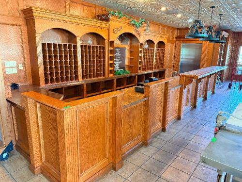 24' LONG CUSTOM-BUILT SOLID OAK BAR AND BACK BAR ASSEMBLY WITH RAISED-PANEL CABINETRY, DECORATIVE MOLDING, CARVED ACCENTS, ARCHED BACK-BAR SECTIONS, MULTIPLE WINE BOTTLE STORAGE CUBBIES, OPEN SHELVING, LOWER STORAGE COMPARTMENTS, SERVICE COUNTER, AND TRIP