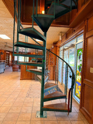 16-STEP METAL SPIRAL STAIRCASE WITH CENTRAL SUPPORT COLUMN, CURVED HANDRAIL, VERTICAL BALUSTERS, AND WOOD-TOPPED METAL TREADS.