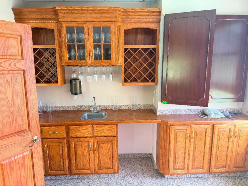 12' CUSTOM OAK WET BAR CABINETRY WITH UPPER AND LOWER CABINETS, BUILT-IN WINE RACKS, GLASS-FRONT DISPLAY CABINET, STEMWARE RACK, WOOD COUNTERTOP, STAINLESS STEEL SINK AND FAUCET. INCLUDES MATCHING BASE CABINET SECTION SHOWN.
