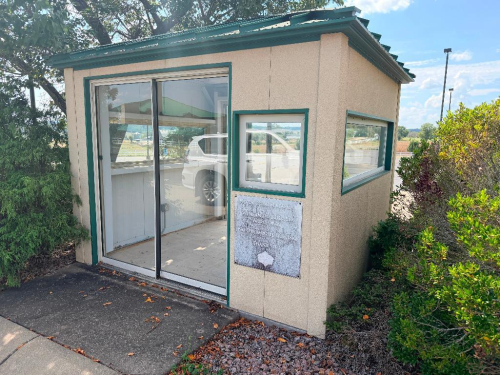 METAL SECURITY / ATTENDANT BOOTH WITH METAL PANEL WALLS AND ROOF, SLIDING GLASS ENTRY DOOR, MULTIPLE WINDOWS, FINISHED INTERIOR WALL PANELS, TILE FLOOR, BUILT-IN METAL DESK, INTERIOR LIGHTING, AND ELECTRICAL OUTLETS.
