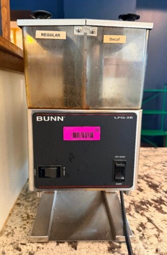 BUNN DUAL-HOPPER COMMERCIAL COFFEE GRINDER WITH TWO SEPARATE BEAN HOPPERS LABELED REGULAR AND DECAF, LEFT/RIGHT HOPPER SELECTOR, START/STOP SWITCH, STAINLESS STEEL HOUSING, AND LOWER BAG RECEIVING PLATFORM.