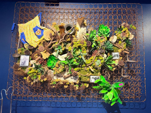 CUSTOM DECORATIVE WALL ART ASSEMBLAGE CONSTRUCTED ON A VINTAGE EXPOSED COIL-SPRING FRAME WITH ARTIFICIAL SUCCULENTS, PRESERVED MOSS, DRIFTWOOD, LOTUS PODS, NATURAL WOOD ELEMENTS, SMALL FRAMED PHOTOGRAPHS, AND ASSORTED DECORATIVE ACCENTS. RUSTED SPRING FRA