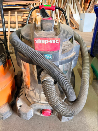 SHOP-VAC HEAVY-DUTY WET/DRY SHOP VACUUM WITH STAINLESS STEEL COLLECTION TANK, LARGE REAR WHEELS, FRONT CASTERS, PUSH HANDLE, DRAIN PORT, AND FLEXIBLE VACUUM HOSE.