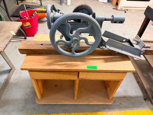 VINTAGE CAST IRON HAND-CRANK MACHINE WITH LARGE FLYWHEEL, GEARED DRIVE ASSEMBLY, ADJUSTABLE FEED MECHANISM AND METAL GUIDE/TRAY, MOUNTED TO WOOD BASE.