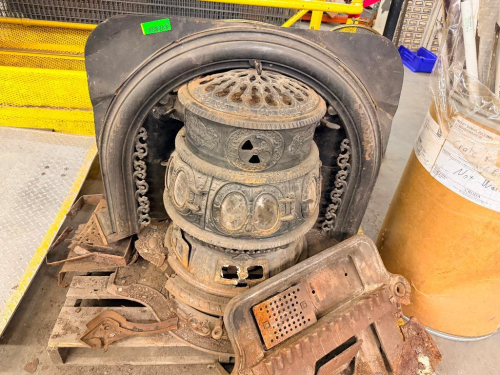 ANTIQUE ORNATE CAST IRON PARLOR STOVE WITH DECORATIVE RELIEF CASTINGS, VENTED DOME TOP, ROUND BODY, CAST BASE AND ASSORTED STOVE COMPONENTS. INCLUDES CAST IRON FIREPLACE SURROUND AND ADDITIONAL LOOSE CAST IRON PARTS.