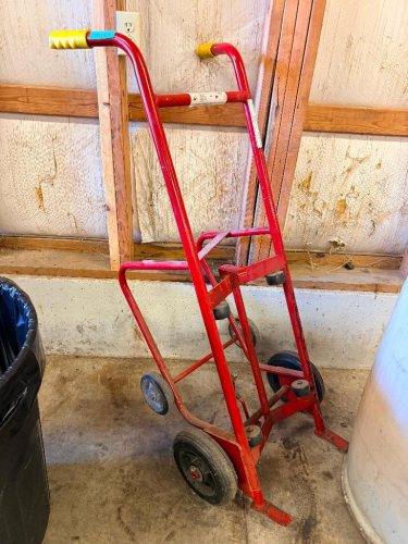 HEAVY-DUTY STEEL DRUM HANDLING DOLLY WITH RED TUBULAR STEEL FRAME, DUAL LARGE RUBBER WHEELS, AUXILIARY SUPPORT WHEELS, RUBBER HANDLE GRIPS AND DRUM ENGAGEMENT BRACKETS.