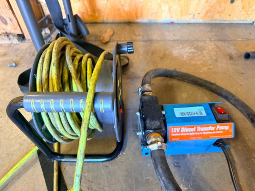 12V DIESEL FUEL TRANSFER PUMP WITH ATTACHED RUBBER HOSES AND PORTABLE ELECTRICAL EXTENSION CORD REEL WITH METAL SUPPORT FRAME.